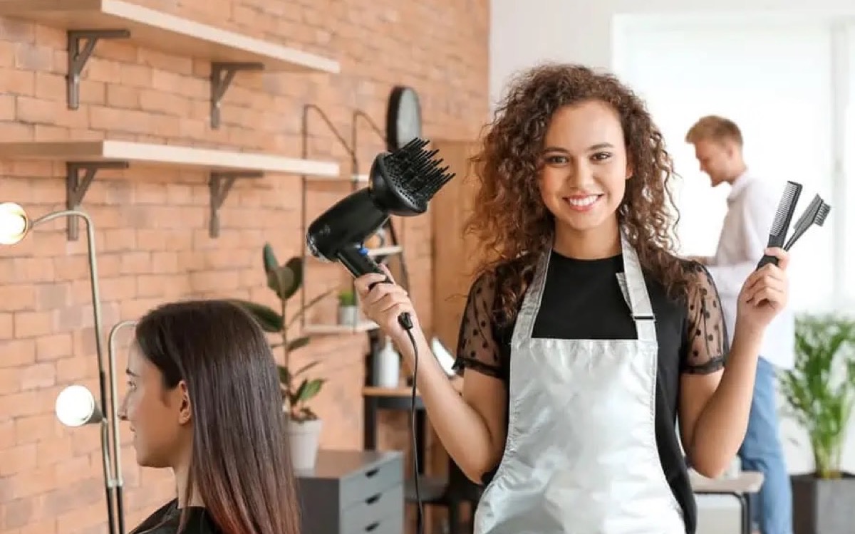 Smiling hairdresser in a modern salon holding a diffuser hair dryer and styling tools in front of a brick wall.