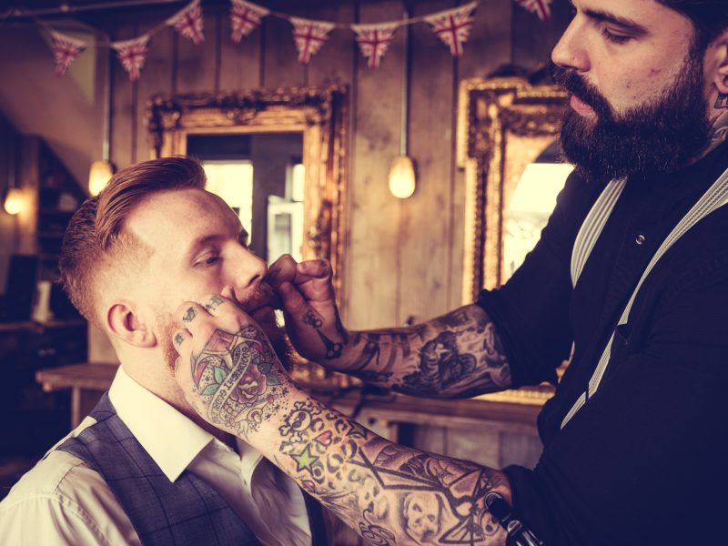 Tattooed barber trimming a client's moustache in a vintage barbershop with warm lighting and Union Jack bunting.